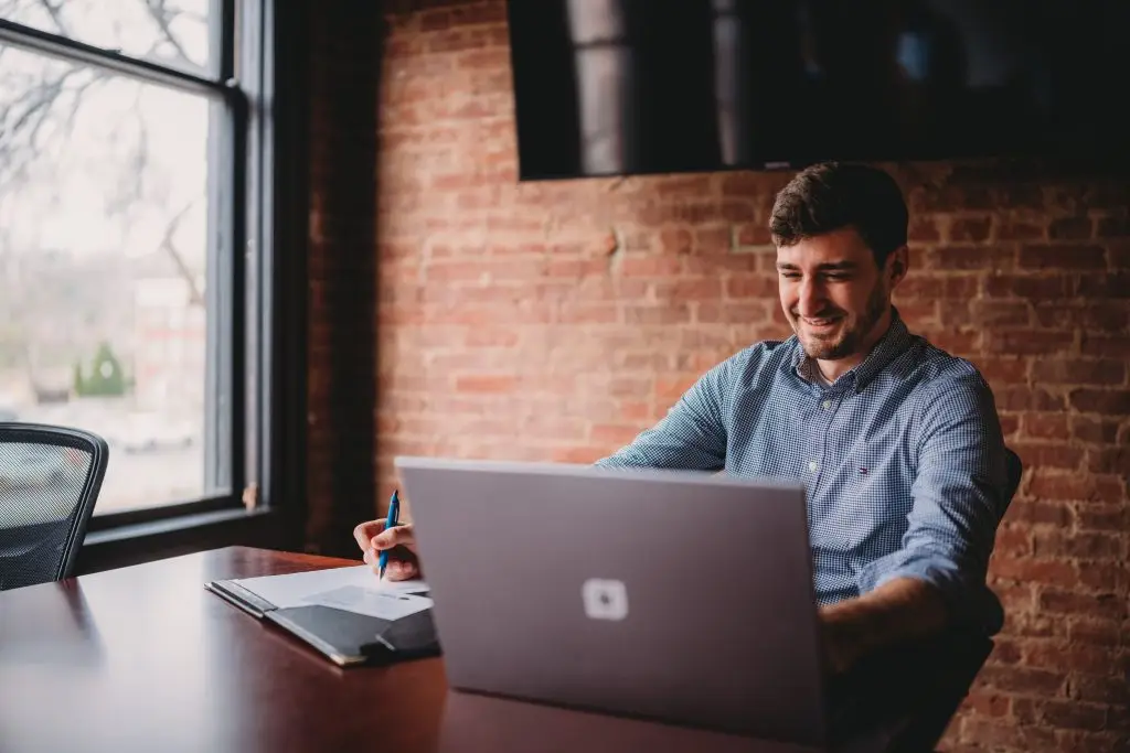 A man sits at a desk using a laptop and a notebook. There is a window and brick wall behind him.