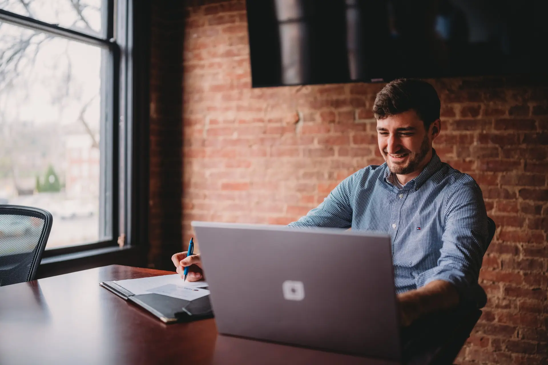 A man sits at a desk using a laptop and a notebook. There is a window and brick wall behind him.